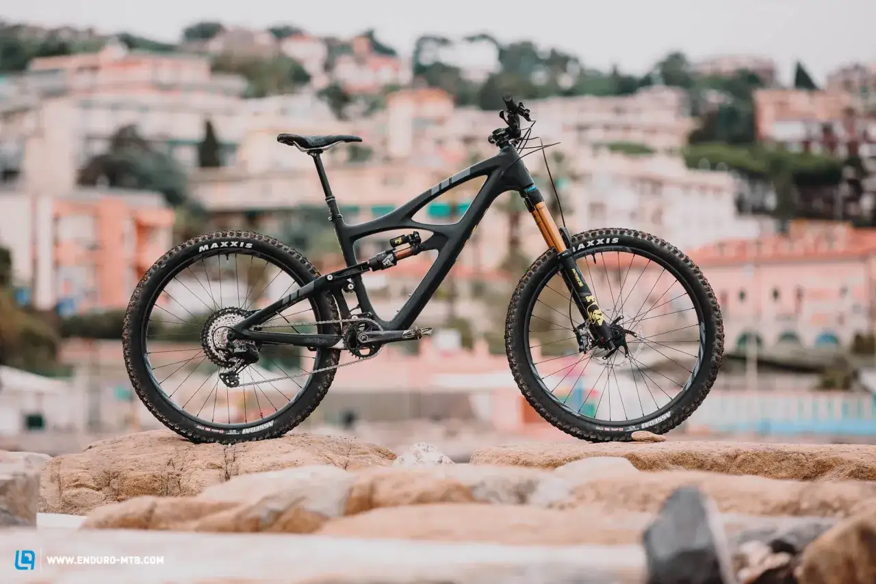 A black Ibis HD5 mountain bike rests on rocks, with a blurred coastal town in the background.