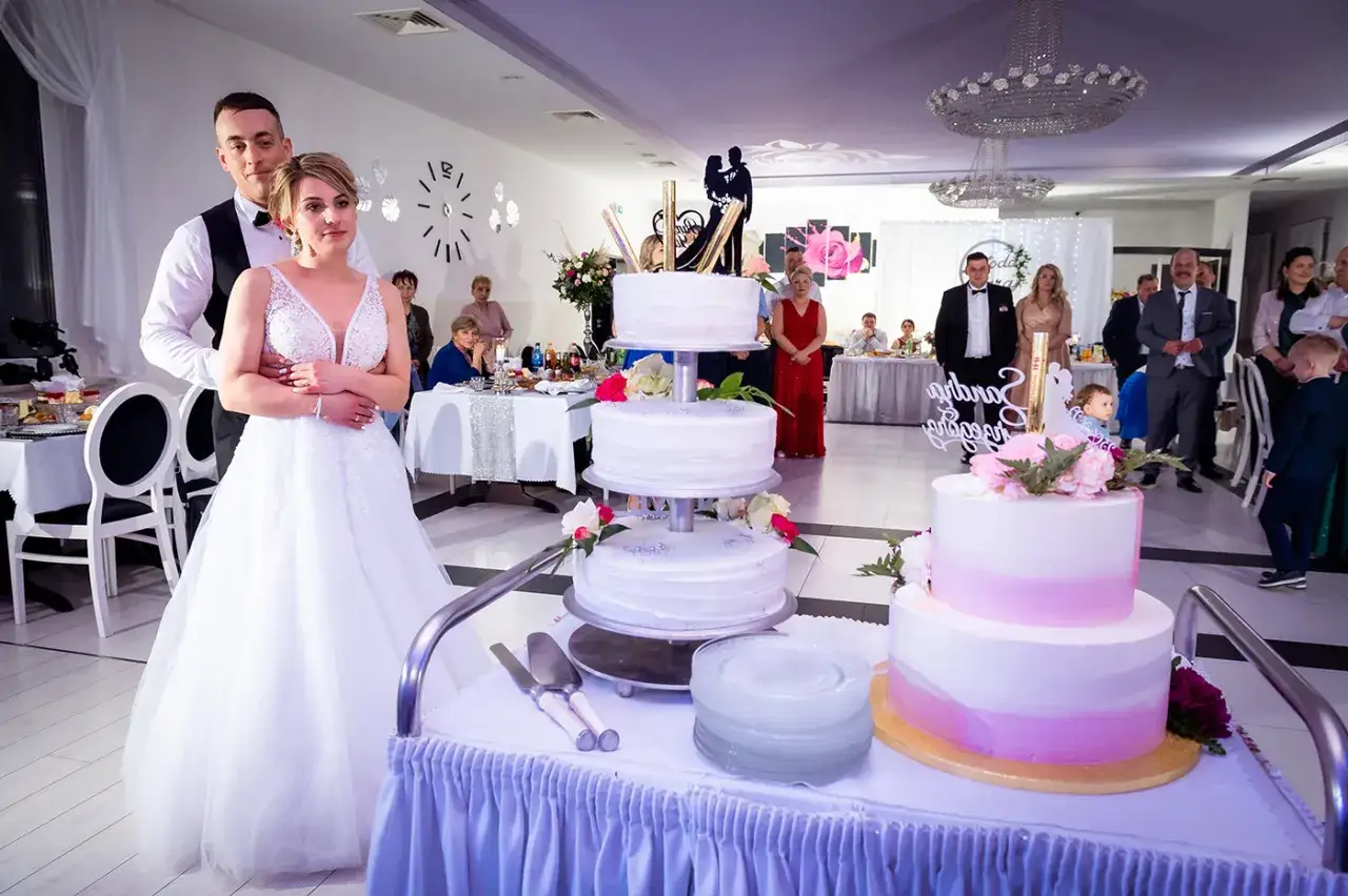 Para 100 osób tort weselny cena jest przystępna. Para młoda pozuje do zdjęcia przy stole z tortami.
