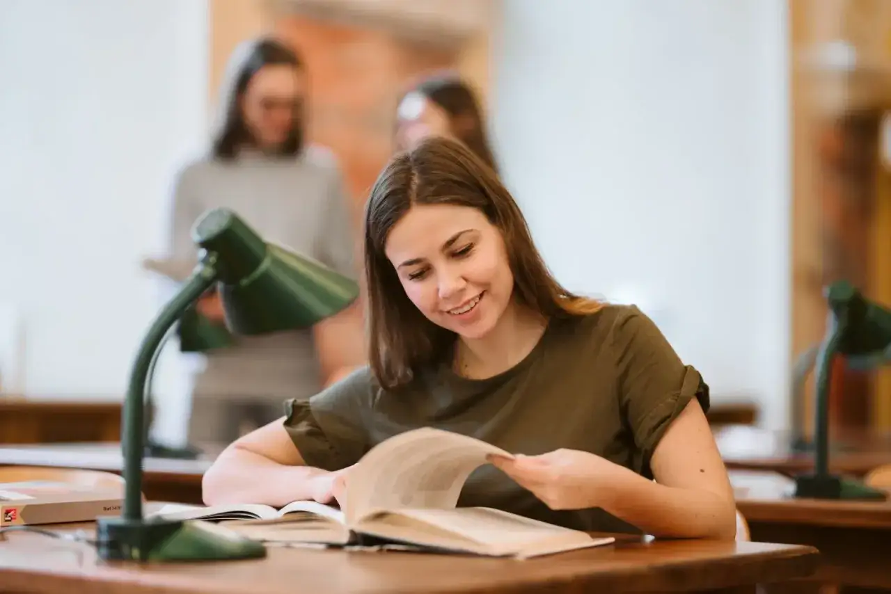 Uśmiechnięta studentka czyta książkę przy lampce. W tle inni studenci. Idealne miejsce na studia coaching Wrocław.
