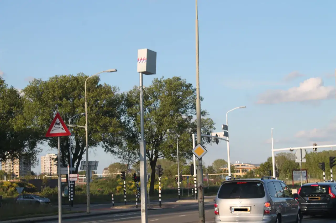 Ein Blitzer in den Niederlanden, auf einem Mast montiert, überwacht den Verkehr. Verkehrsschilder warnen vor rutschiger Fahrbahn.