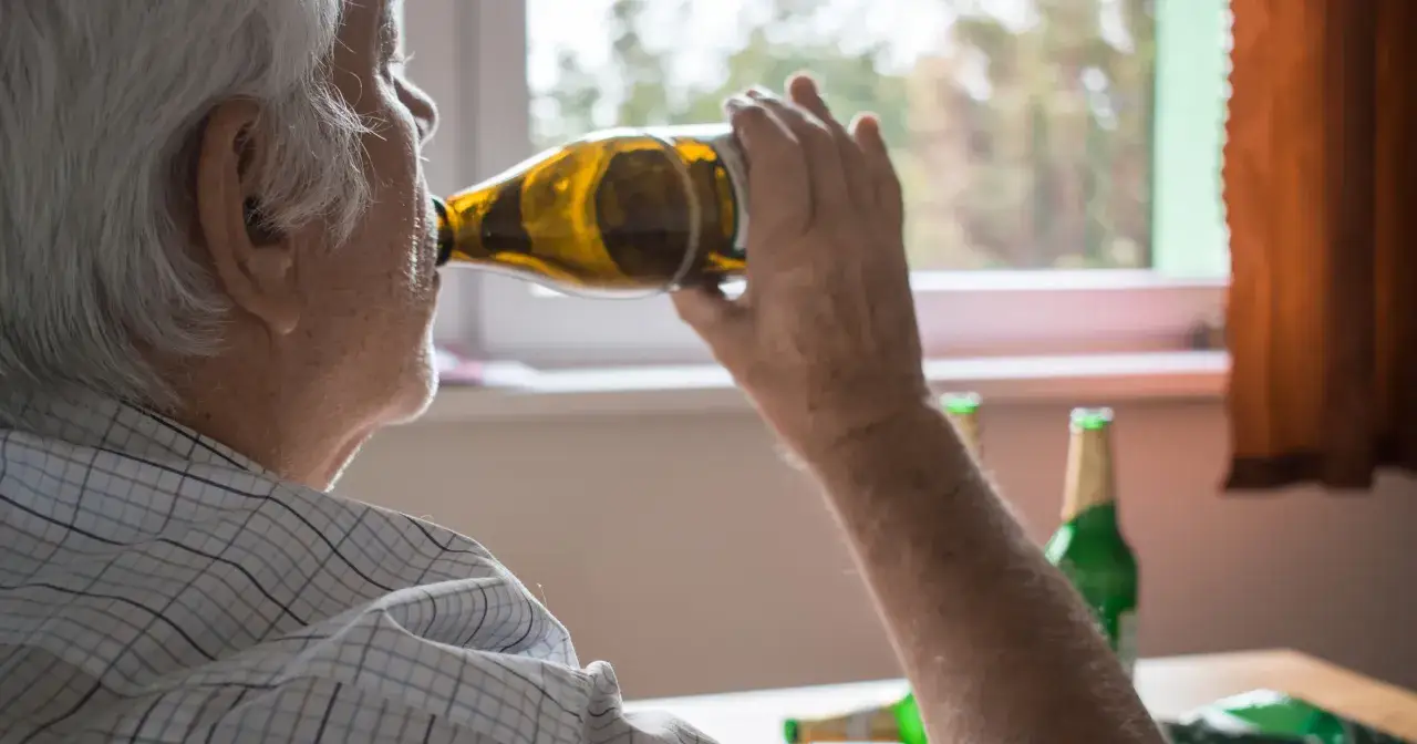 Starszy mężczyzna pije piwo z butelki. W tle widać inne butelki. Zastanawiasz się, renta alkoholowa ile wynosi?