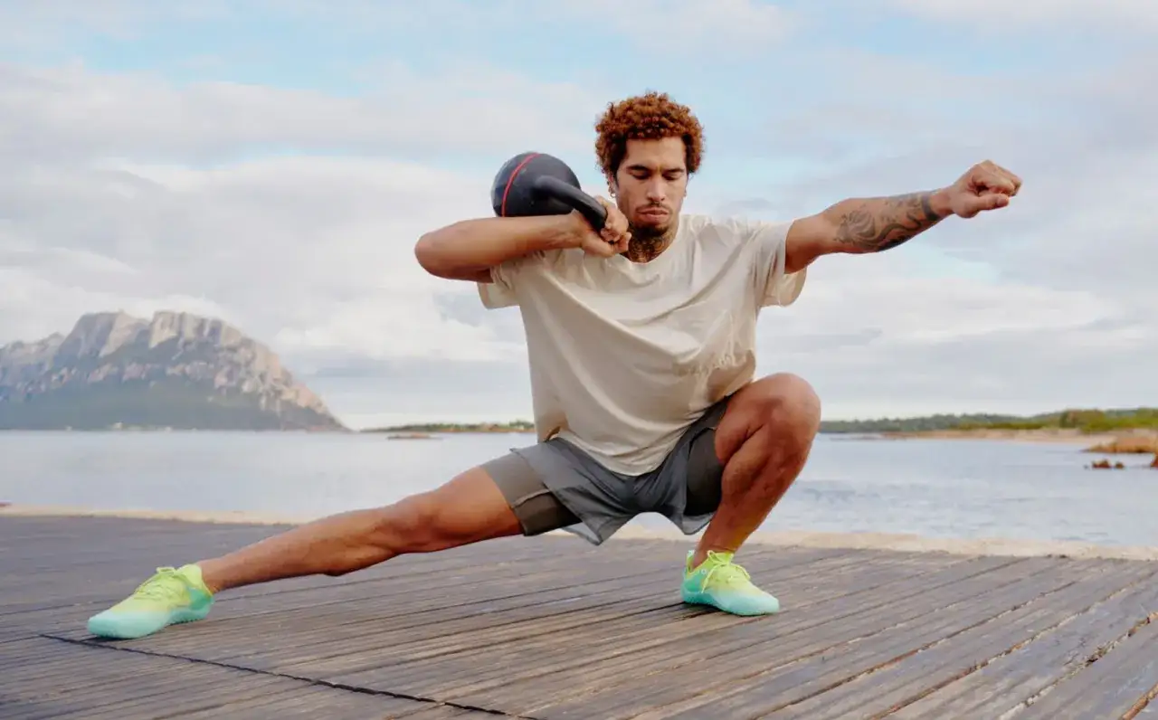 Mann trainiert mit Kettlebell am Meer, seitliche Ausfallschritte, **ohne schuhe** auf Holzplanken.