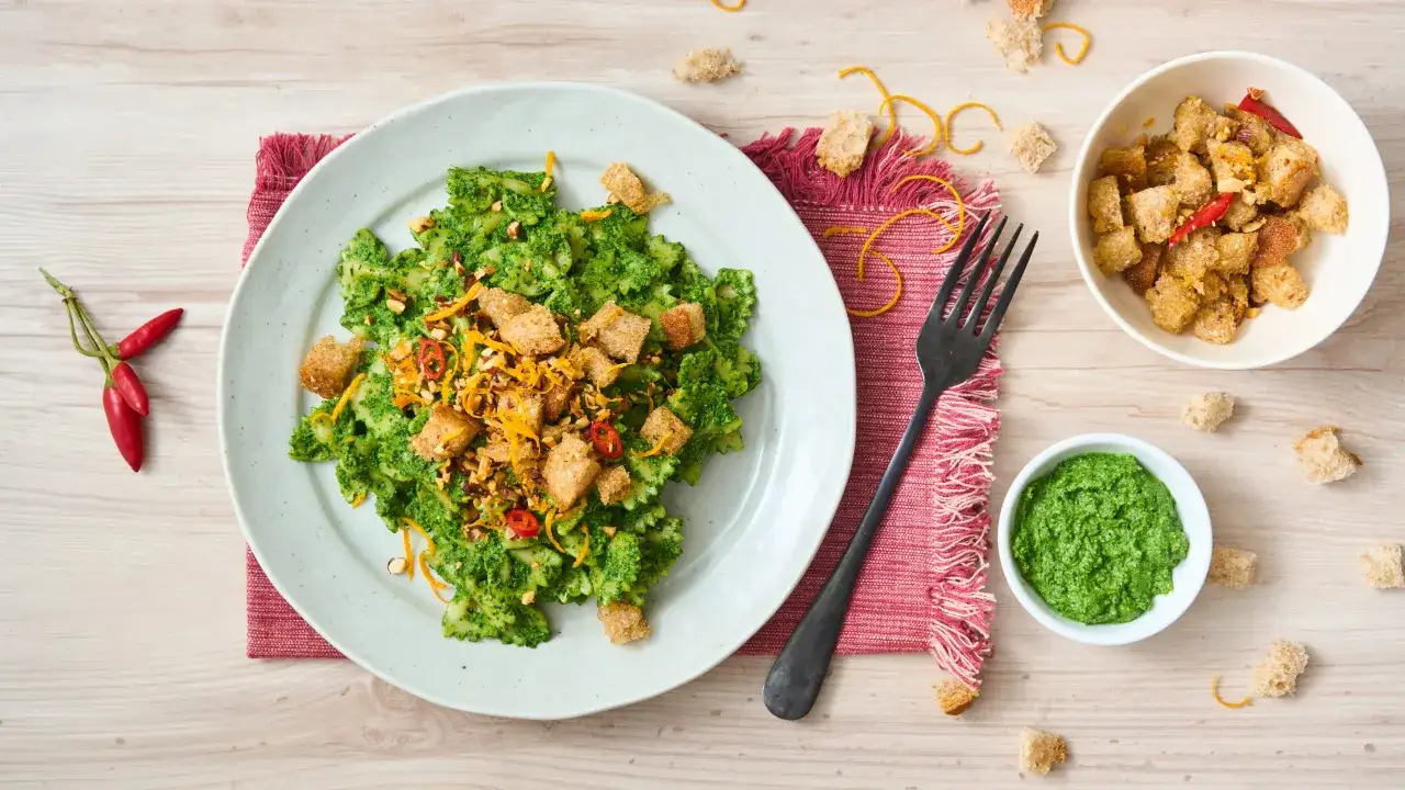 Farfalle condite con pesto di cavolo nero, crostini, peperoncino e scorza d'arancia. Accanto, una ciotolina di pesto e ingredienti sparsi.