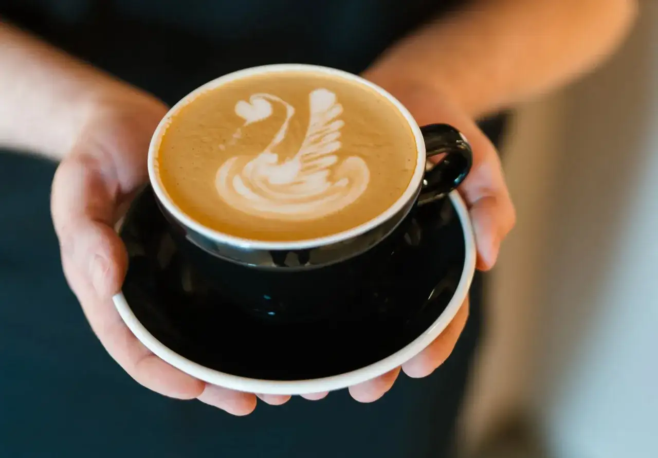 Dłonie trzymają czarną filiżankę z kawą latte z pianką w kształcie łabędzia. Zastanawiasz się, ile kalorii ma kawa latte?