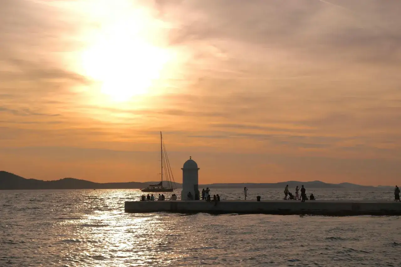 Zach&oacute;d słońca nad Zadar, z ludźmi spacerującymi po molo i żagl&oacute;wką na morzu. Czy warto? Zdecydowanie tak!