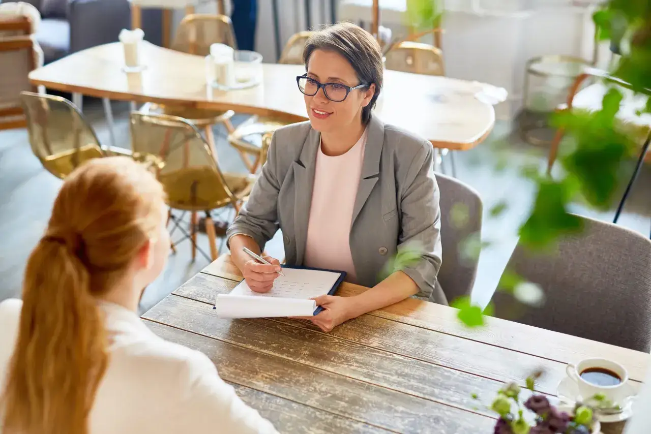 Entrevista de seleccion personal hosteleria. Una mujer con gafas revisa un documento en una mesa de madera, mientras otra persona, de espaldas, escucha atentamente.
