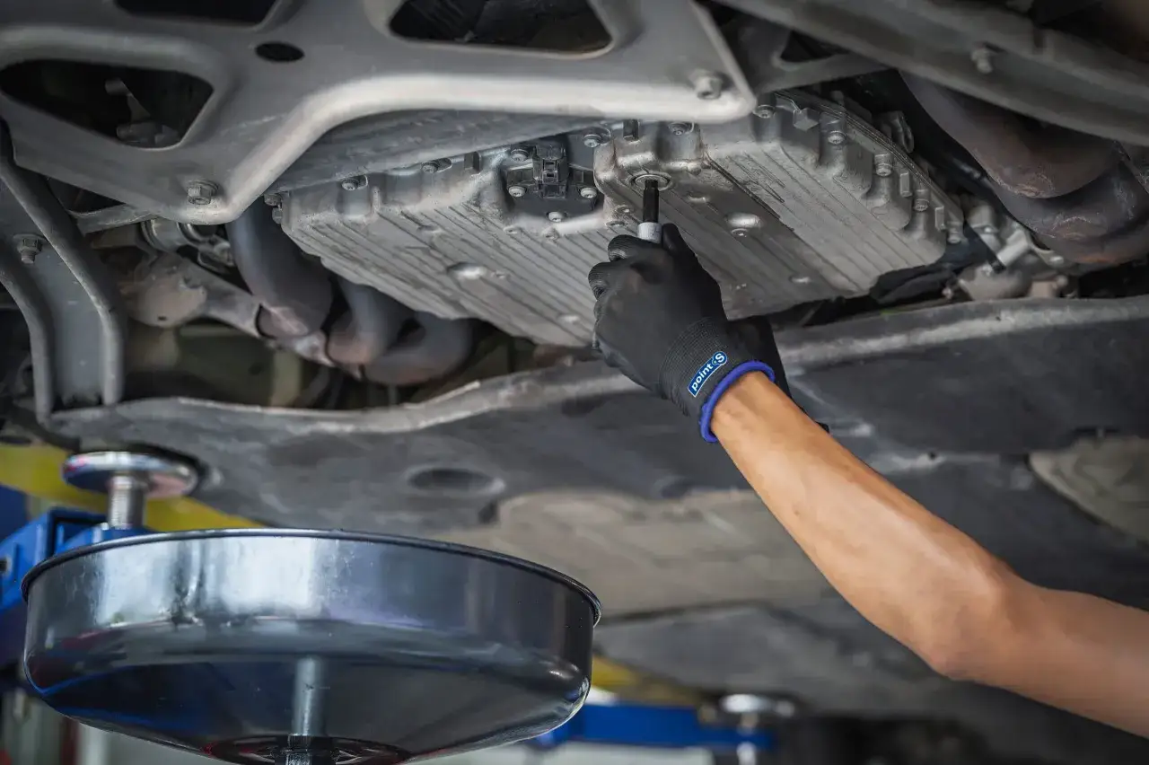 Main d'un mécanicien en gant noir dévissant le bouchon de vidange d'une voiture diesel, avec un bac de récupération d'huile en dessous.