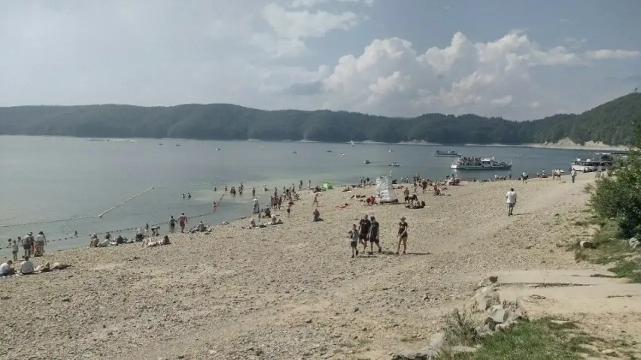 Plaża nad Soliną pełna ludzi, którzy korzystają z kąpieli i słońca. Widać łodzie i statki wycieczkowe.