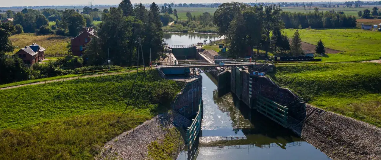 Widok na śluzę gdańską głowę, otoczoną zielenią i budynkami. Woda w kanałach odbija niebo.