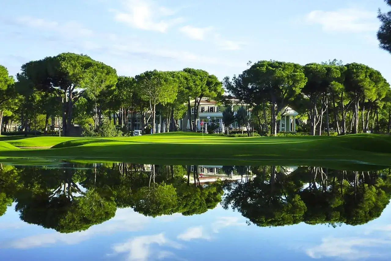Grüner Golfplatz mit einem Gebäude im Hintergrund, umgeben von Bäumen. Das Wasser spiegelt die Szene wider, wie bei Robinson Nobilis.