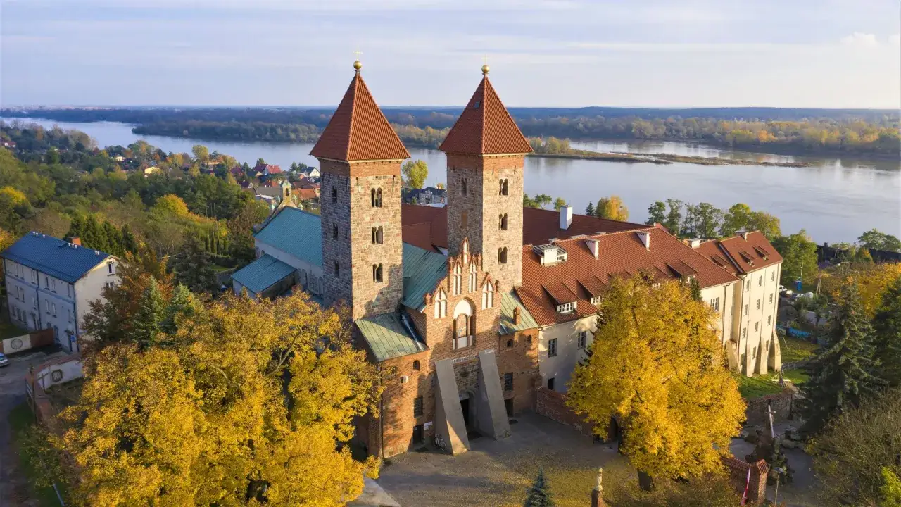 Dwukopułowa bazylika w Czerwińsku nad Wisłą, otoczona jesienną zielenią, z szeroką rzeką w tle.