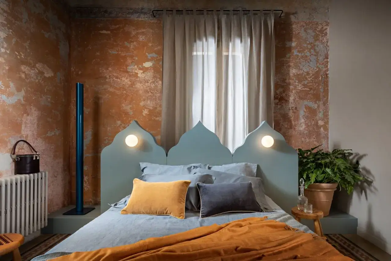 Camera da letto con testiera azzurra a forma di arco, cuscini color ocra e antracite, coperta arancione. Parete con intonaco a vista, lampada blu. Ispirazioni per un ambiente unico.