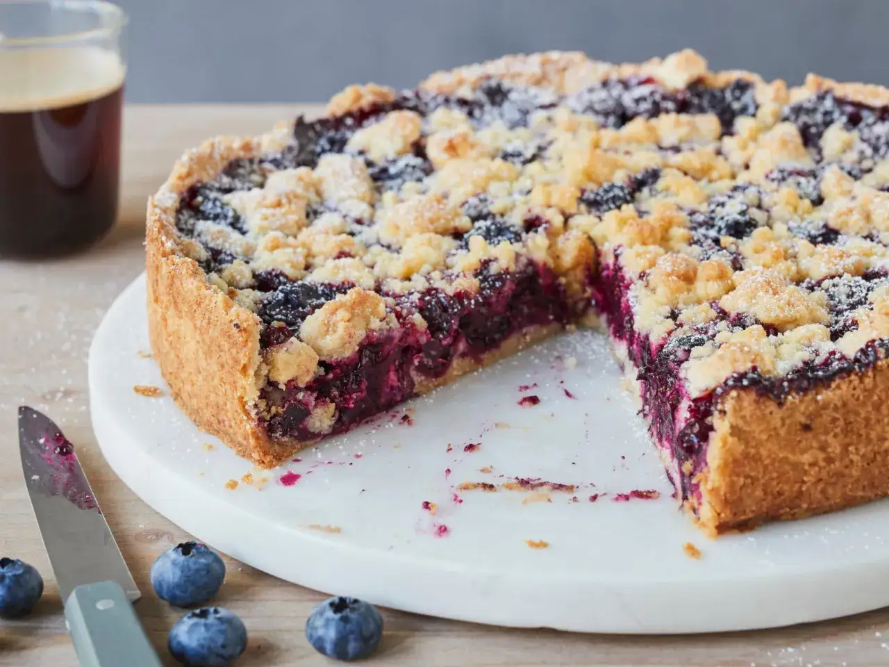 Ein saftiger Heidelbeerkuchen Landfrauenart mit Streuseln, daneben frische Heidelbeeren und ein Glas Kaffee.