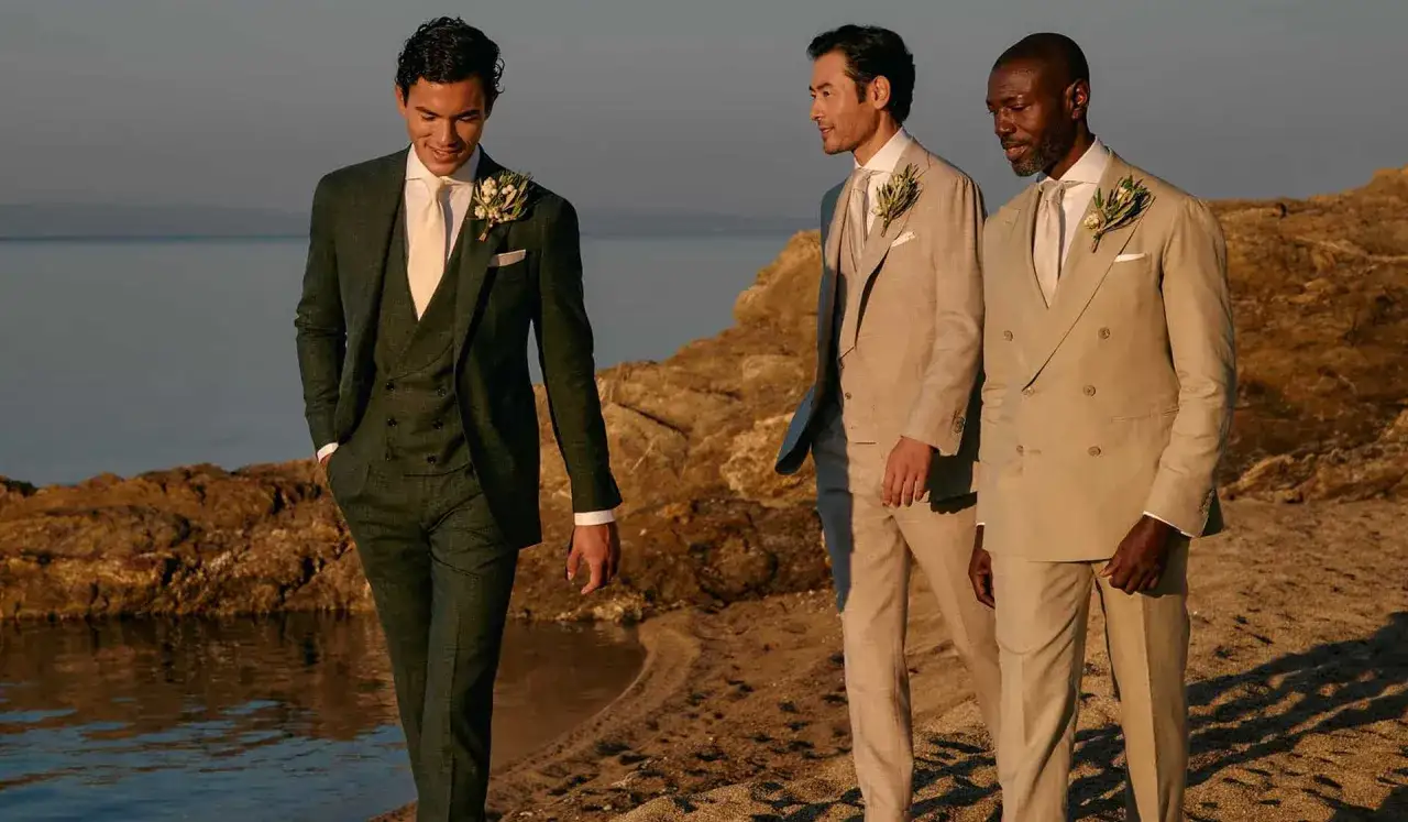 Three men in stylish suits, perfect for a black tie wedding guest, walk on a beach by the sea.