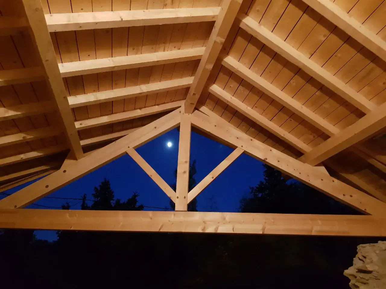 Vue sous un ciel étoilé, la charpente traditionnelle 2 pans en bois clair, avec ses poutres robustes, offre un aperçu du ciel nocturne et de la lune.
