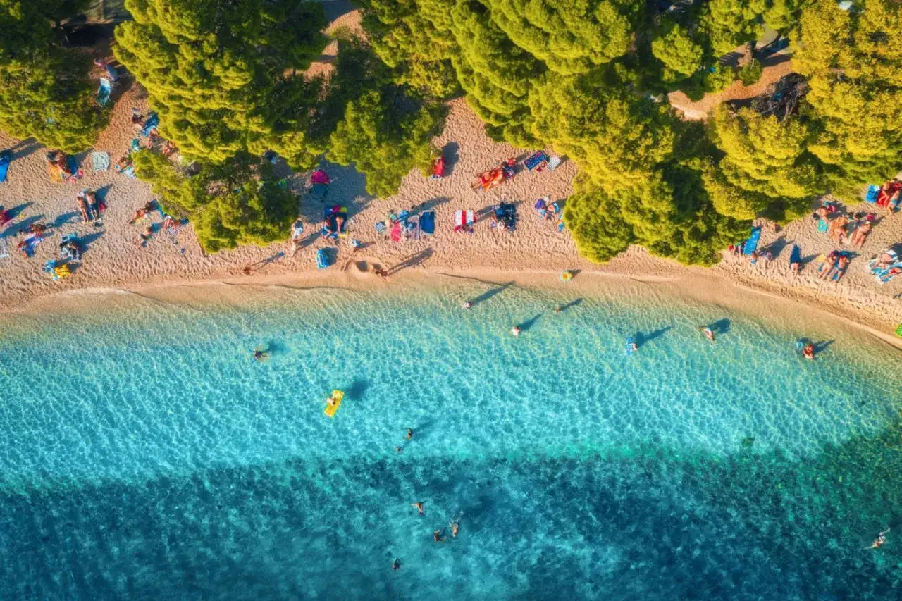 Słoneczne plaże w Chorwacji pełne ludzi odpoczywających pod drzewami i kąpiących się w krystalicznie czystej wodzie.