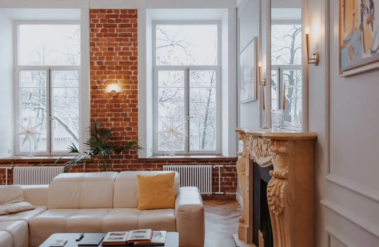 Przytulny salon z białą kanapą, kominkiem i oknami wychodzącymi na zimowy ogród. Ciepła ściana z cegły aranżacje dodaje wnętrzu charakteru.