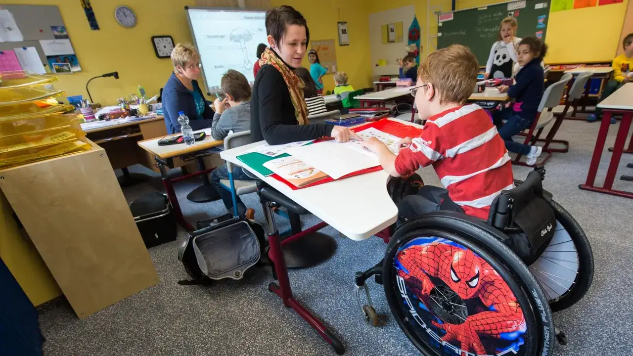 Lehrerin unterstützt Jungen im Rollstuhl beim Lernen. Ein Beispiel für gelebte **Inklusion an Schulen**, wo alle Kinder gemeinsam lernen.