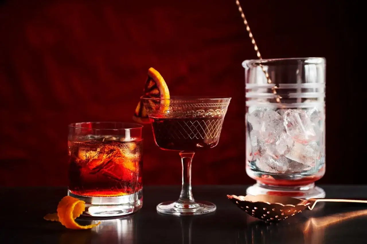 Drei Gläser mit roten Cocktails, Eiswürfel, ein Glas mit Eis und ein Kupfer-Barsieb. Die Zutaten für einen Negroni sind bereit.