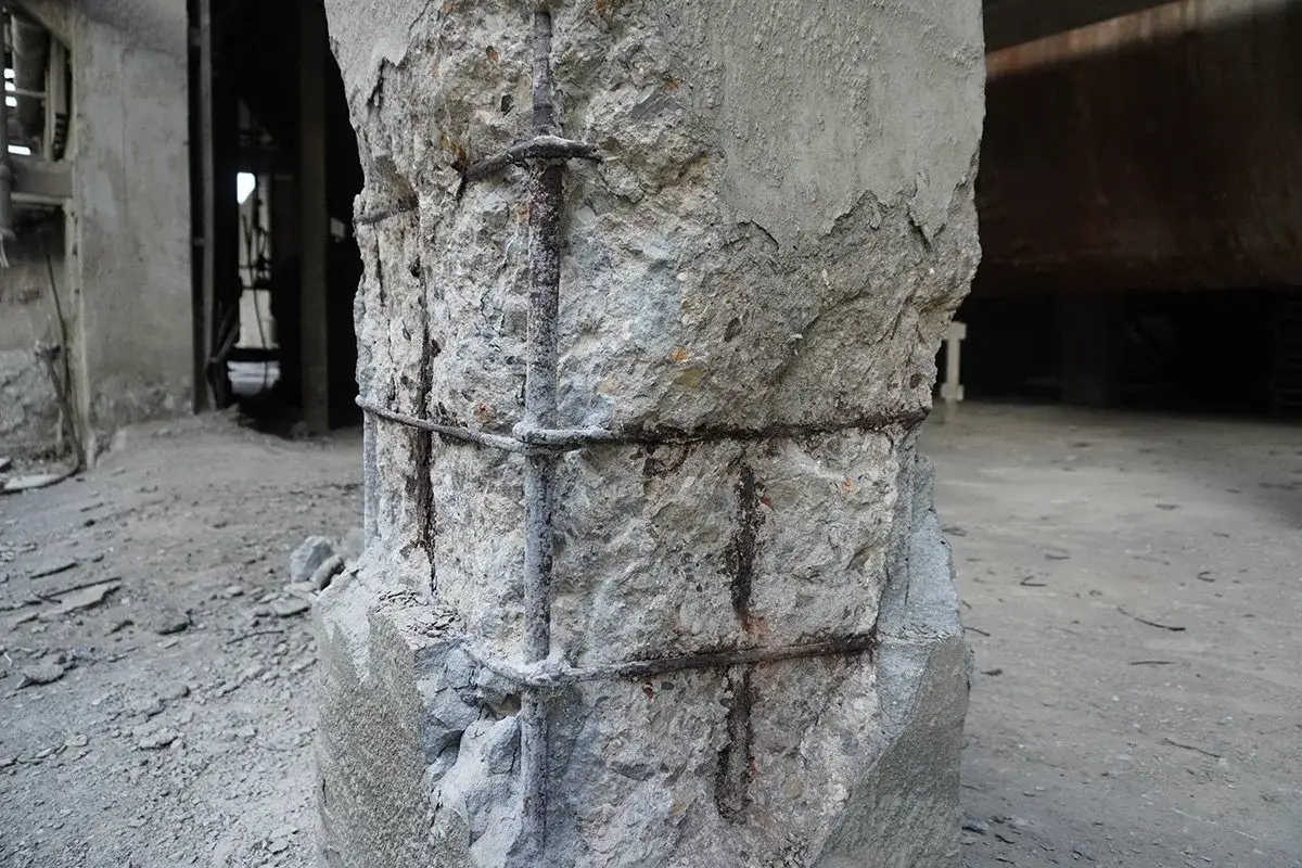 Columna de hormig&oacute;n fraguado r&aacute;pido con barras de refuerzo expuestas y corro&iacute;das, mostrando signos de deterioro en un entorno industrial abandonado.