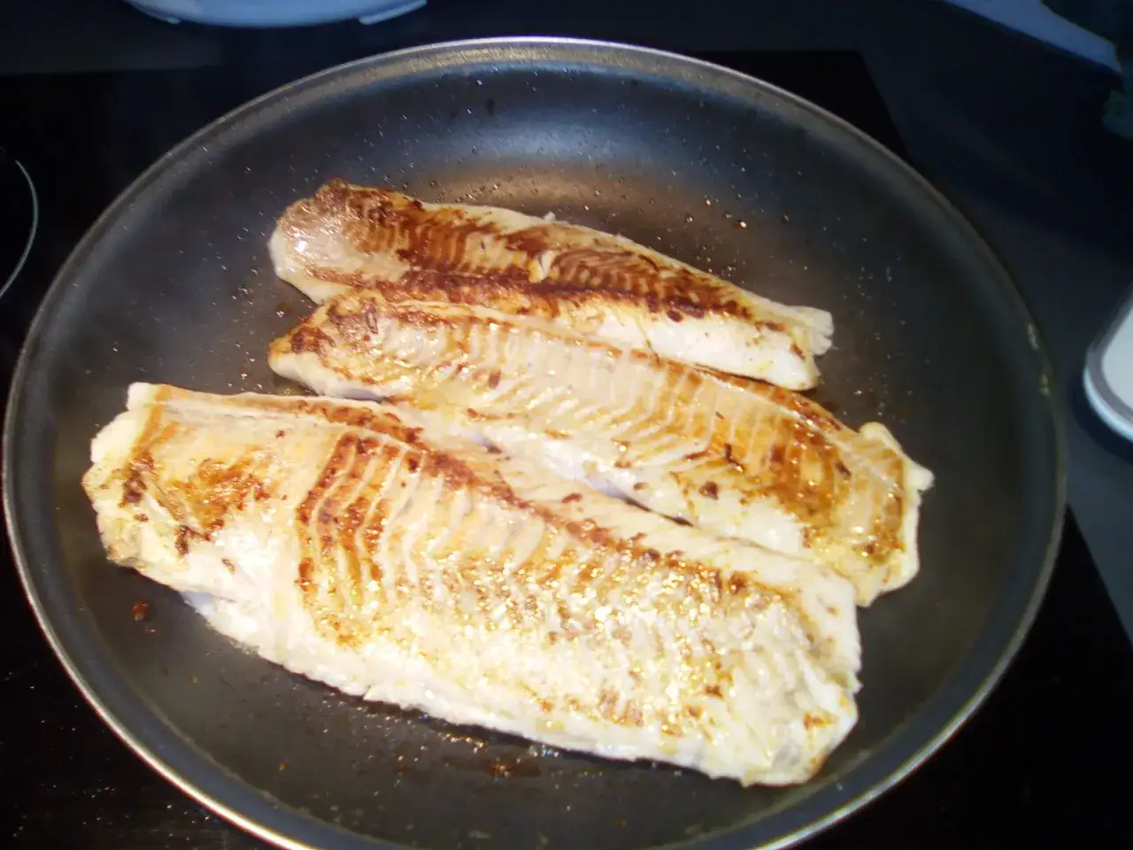 Trois filets de lieu noir dorent dans une poêle, leur chair blanche contrastant avec les marques de cuisson dorées.