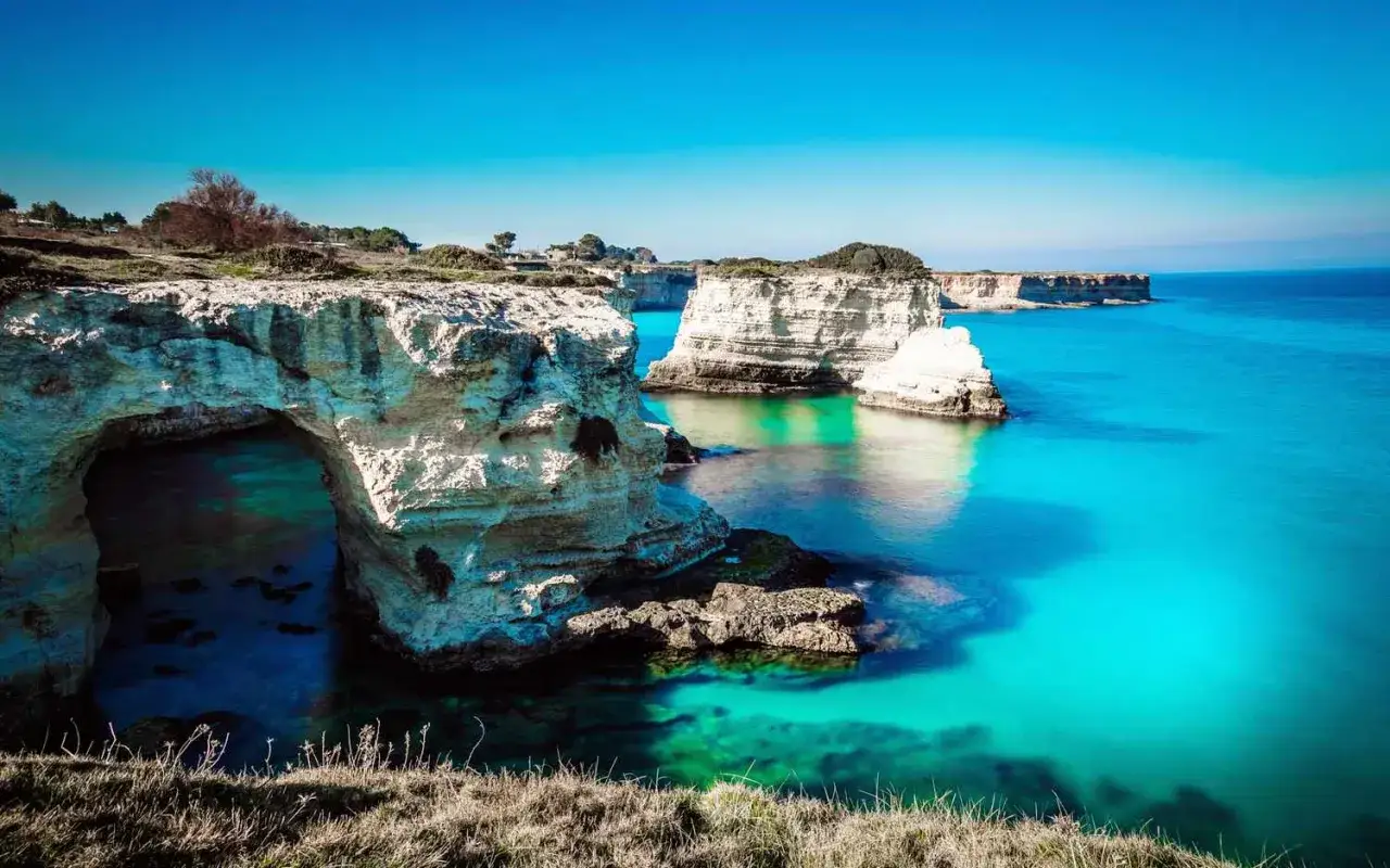 Klippor och turkost vatten vid kusten i Salento, Italien. En naturlig stenbro bildar en ingång till en grotta.