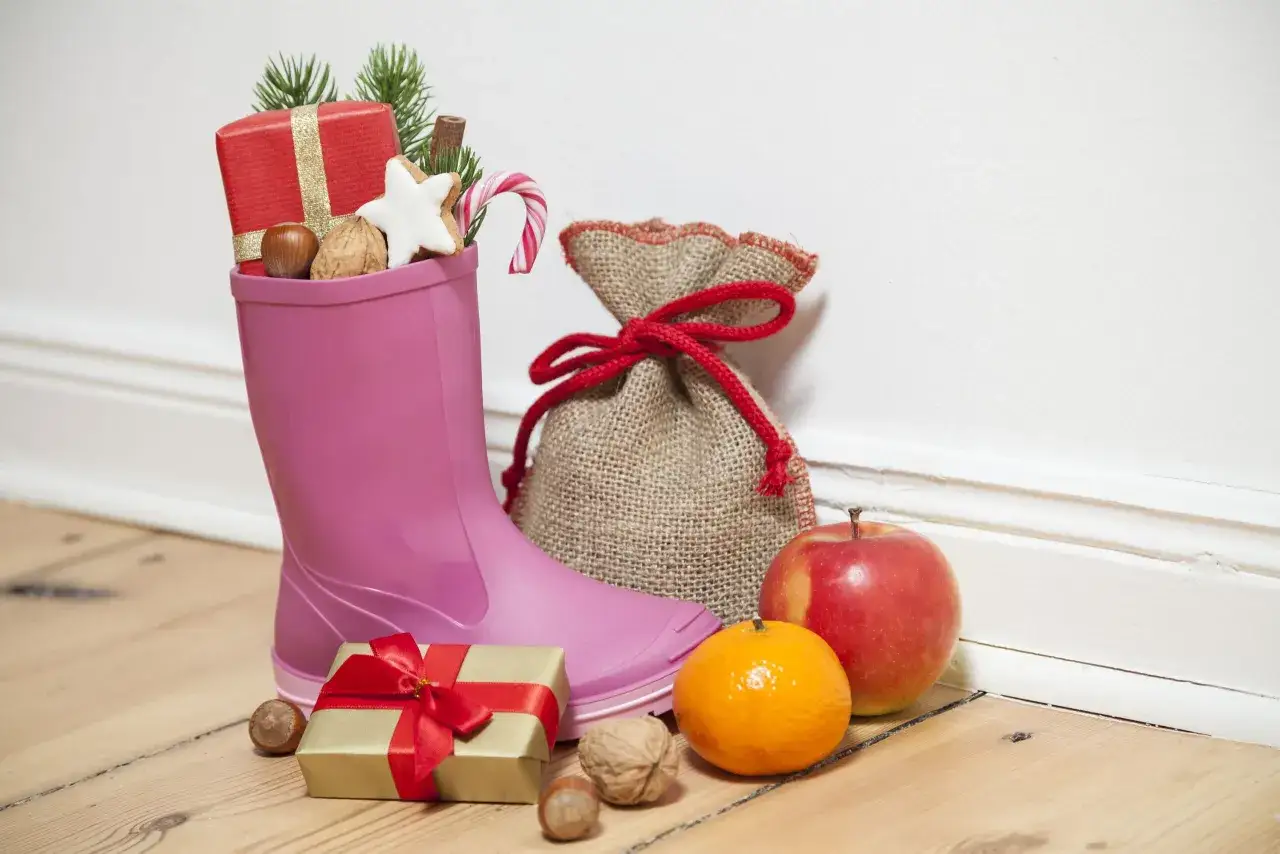 Eine pinke Gummistiefel gefüllt mit Geschenken, Nüssen und Süßigkeiten, bereit für den Nikolaus. Daneben ein Jutesack, ein Geschenk, Mandarine und Apfel.