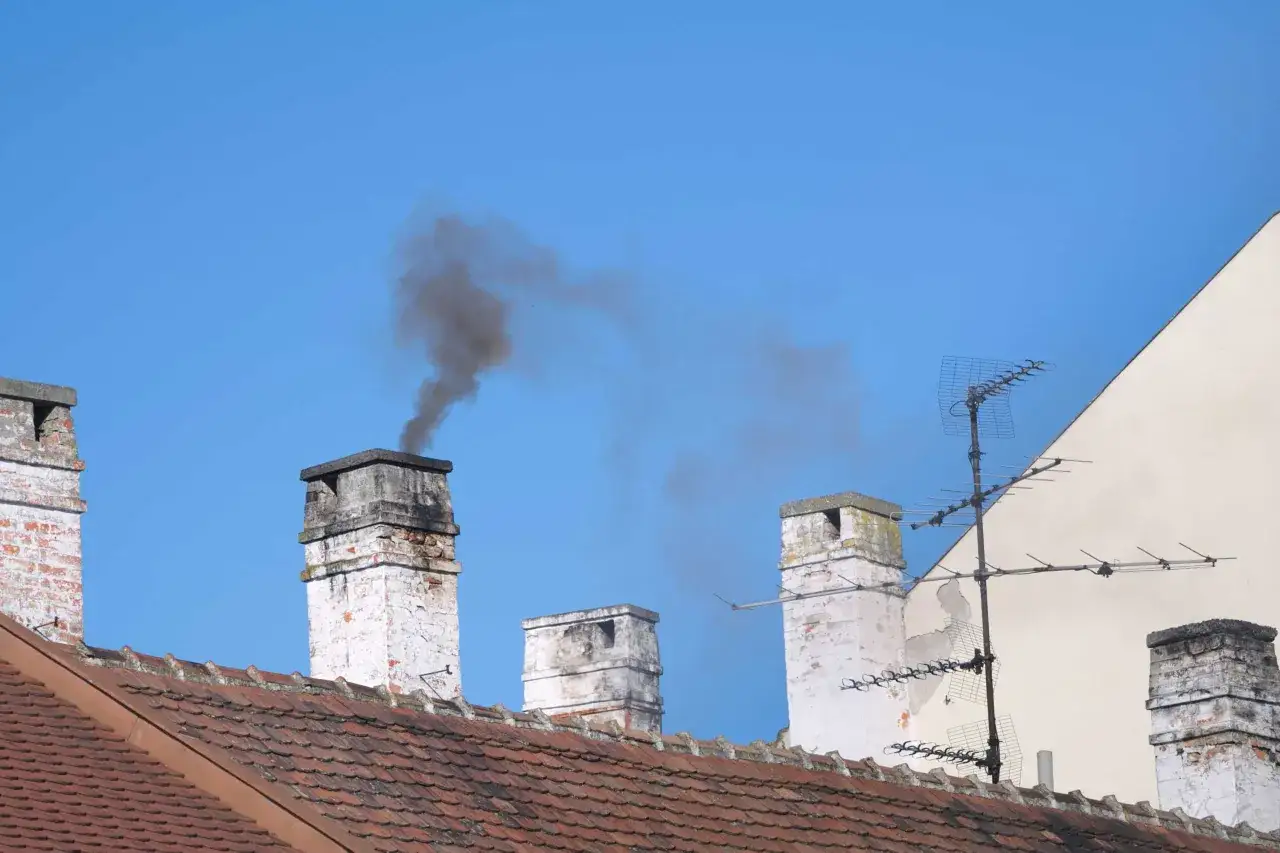 Fumée noire s'échappant d'une cheminée sans tubage, sur fond de ciel bleu. Plusieurs cheminées et une antenne TV sont visibles sur le toit.