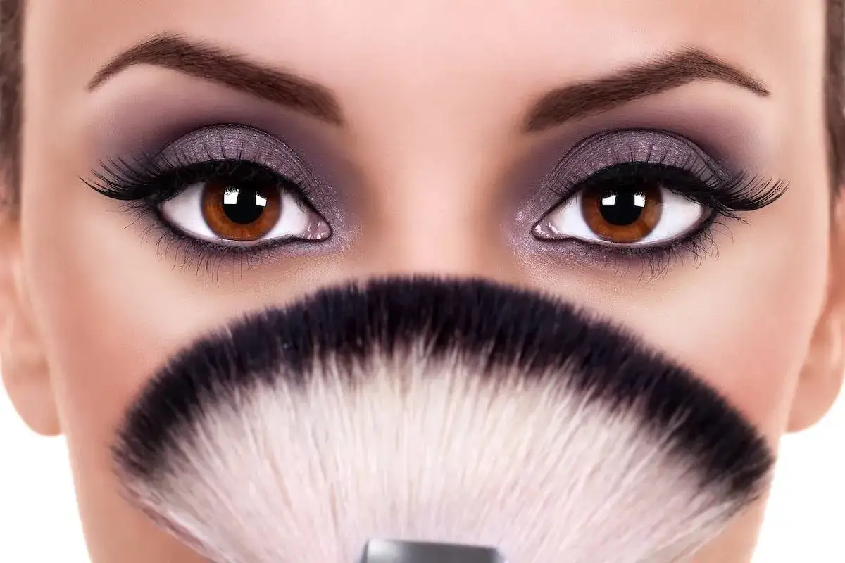 Occhi grandi e tondi esaltati da un trucco smokey viola e ciglia finte, con un pennello da trucco in primo piano.