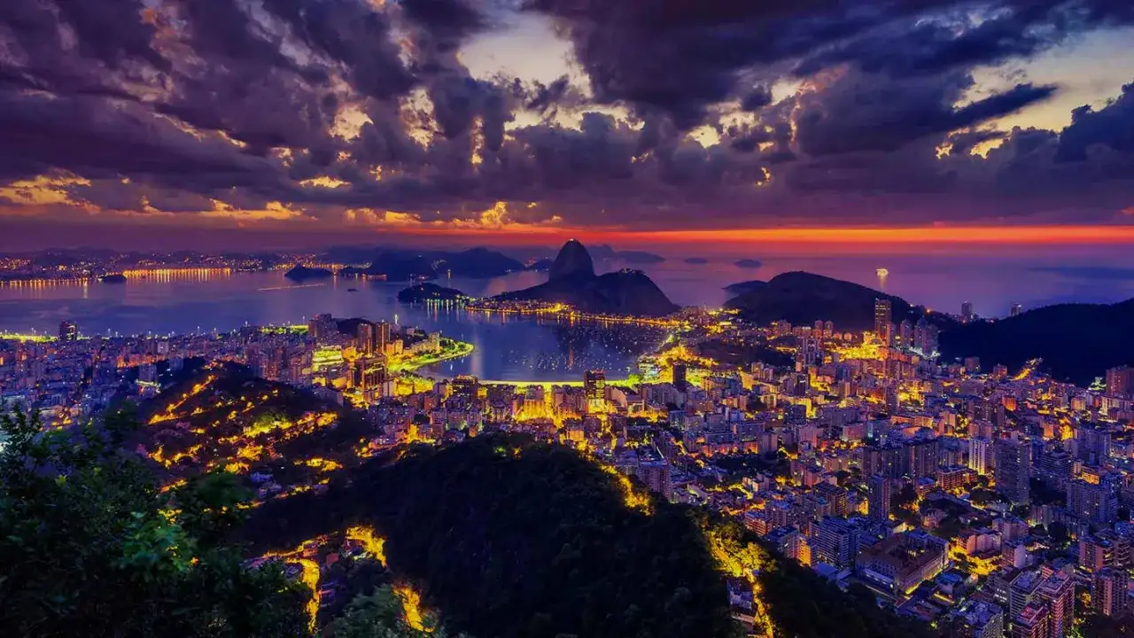 En hisnande vy över Rio de Janeiro i skymningen. Staden lyser upp med tusentals ljus, medan himlen färgas i lila och orange. Sockertoppen reser sig majestätiskt i bakgrunden.