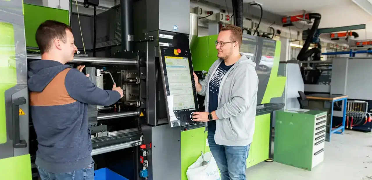 Zwei M&auml;nner bedienen eine Spritzgussmaschine. Einer stellt die Maschine ein, der andere bedient das Touchpanel. Perfekt f&uuml;r ein Studium im Maschinenbau berufsbegleitend.
