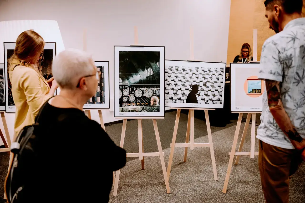 Ludzie oglądają prace na konkursie fotograficznym dla amatorów. Wystawa prezentuje ciekawe ujęcia, od zegarów po architekturę.