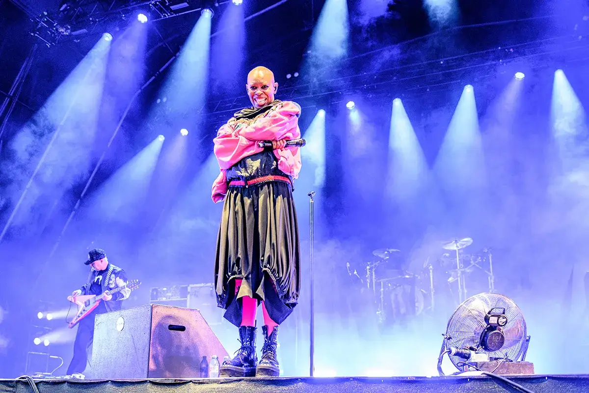 Skunk Anansie na żywo! Wokalistka Skin w różowej bluzie i czarnej spódnicy, z mikrofonem w dłoni, na tle niebieskich świateł i dymu.