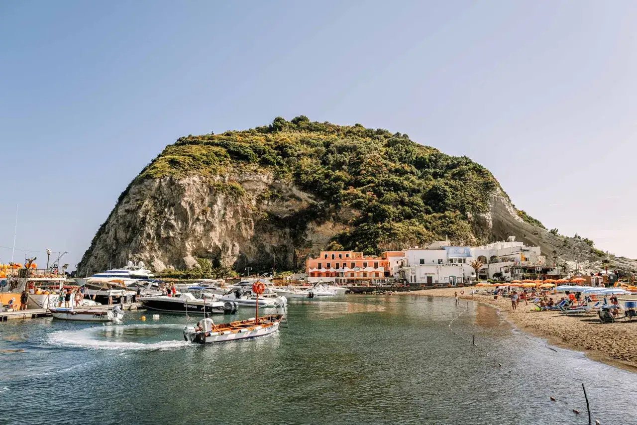 La marina di Sant'Angelo, con le sue barche ormeggiate e la spiaggia affollata, si estende ai piedi di un'imponente collina verdeggiante.