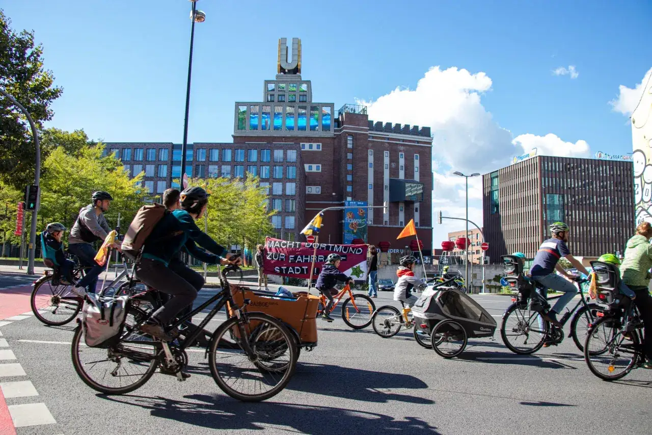 Fahrradfahrer, darunter Kinder, nehmen an der Kidical Mass Dortmund teil. Sie fahren an Geb&auml;uden vorbei, auf denen 
