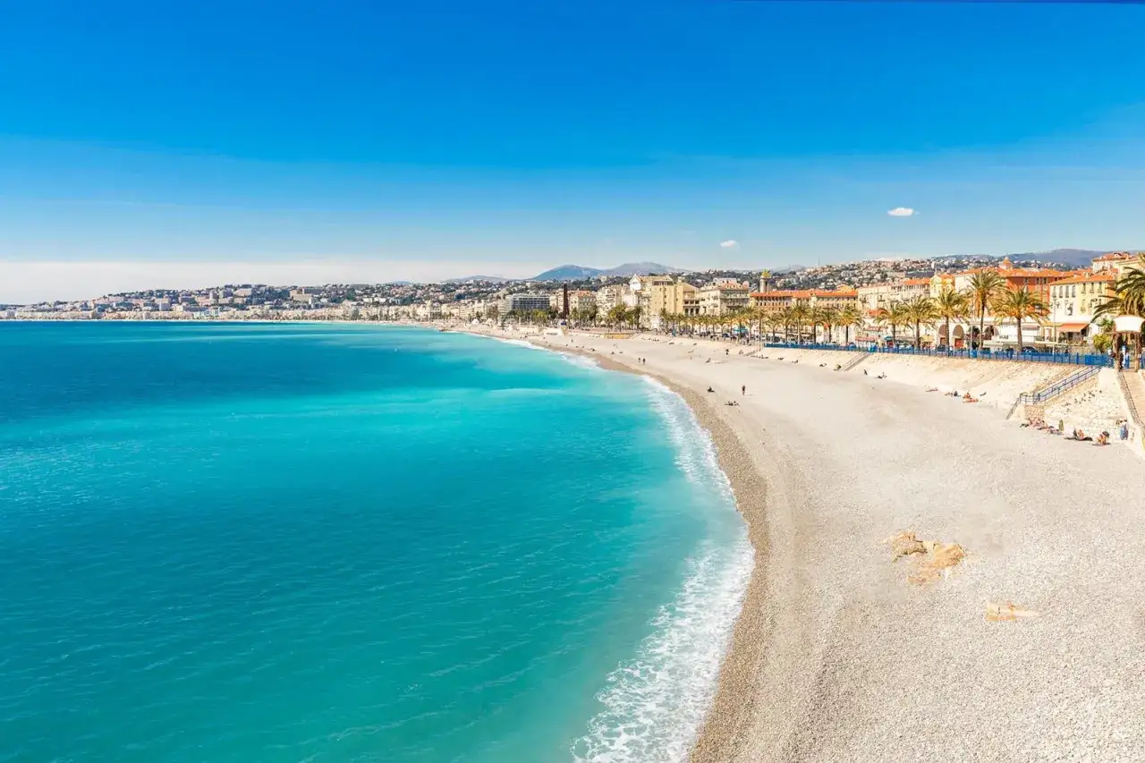 Spiaggia di ciottoli baciata dal sole sulla costa azzurra, con il mare turchese che lambisce la riva e la città sullo sfondo.