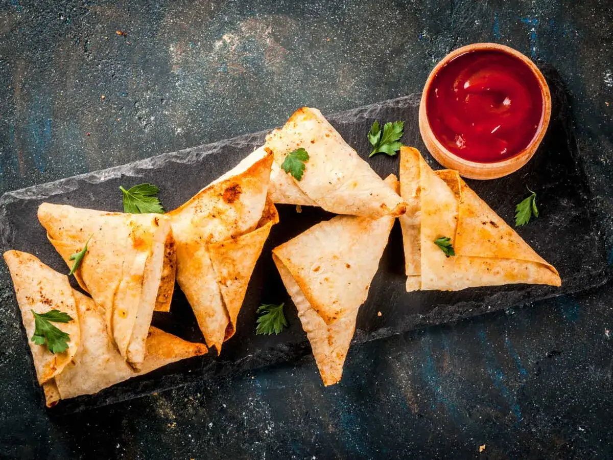 Des samoussas poulet dor&eacute;s et croustillants, parsem&eacute;s de persil frais, accompagn&eacute;s d'un bol de sauce tomate sur une planche d'ardoise.