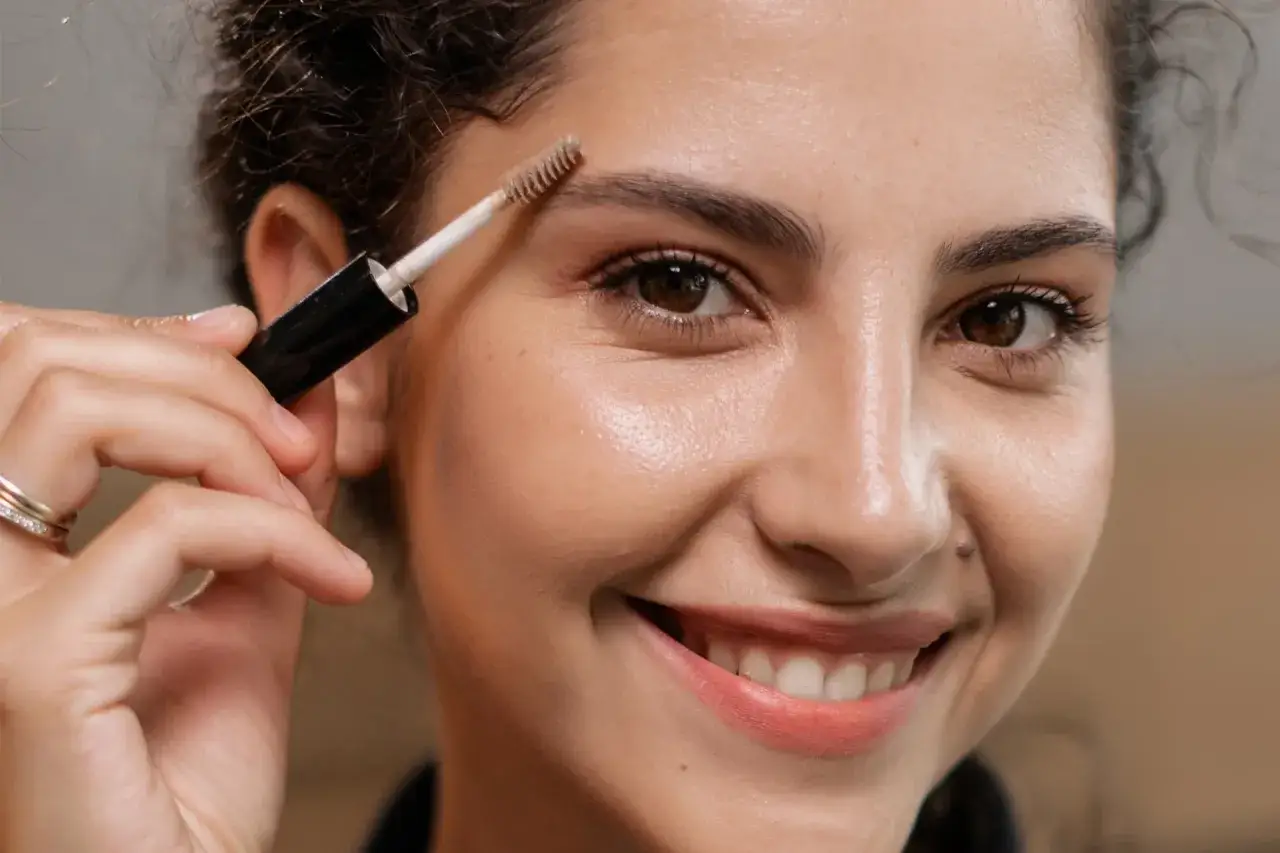 Mujer sonríe mientras se arregla la **ceja** con un cepillo de maquillaje.