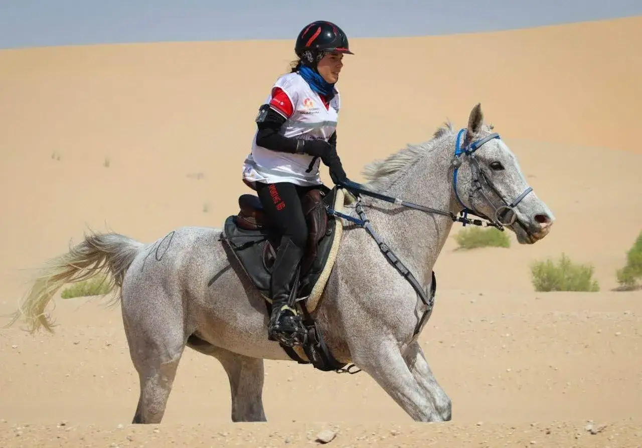 Une cavalière et son pur sang arabe endurance traversent le désert. Le cheval gris moucheté court sur le sable, la cavalière porte un casque et une tenue de compétition.