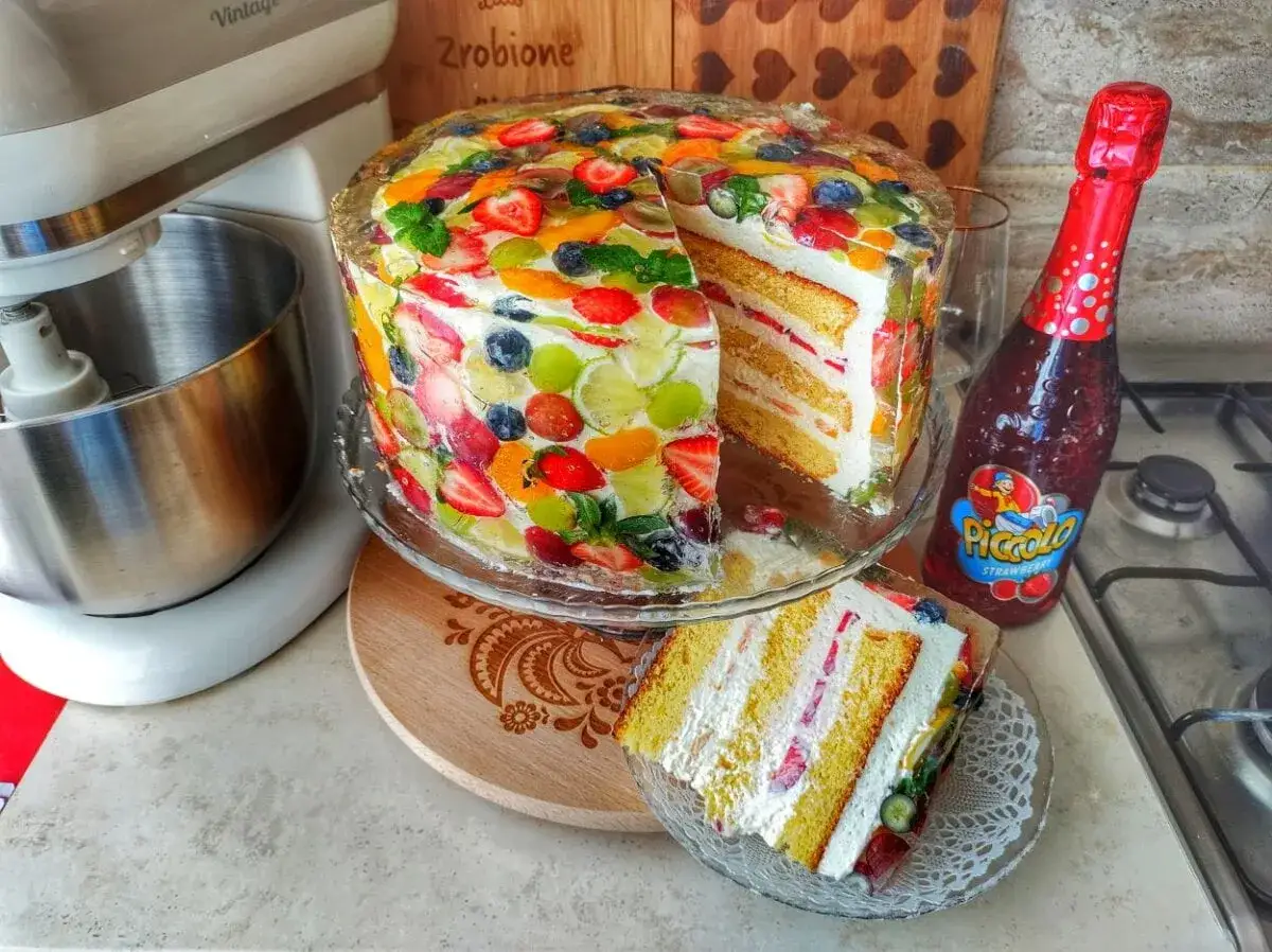Kolorowy przekr&oacute;j tortu z galaretką i owocami, obok butelka Piccolo.
