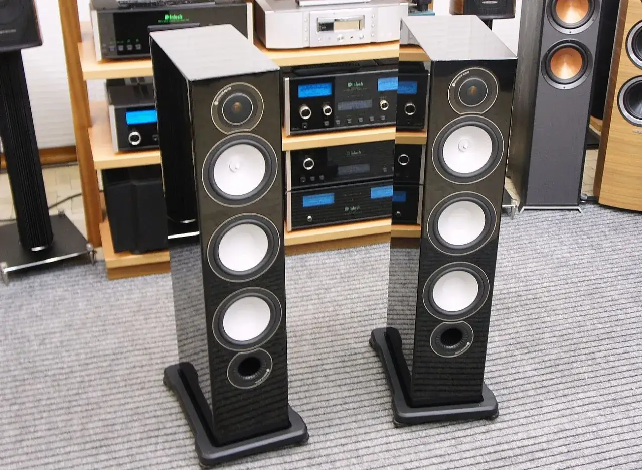 Dwie eleganckie kolumny głośnikowe Monitor Audio RX8 w czarnym połysku, stojące na szarym dywanie, obok sprzętu audio.