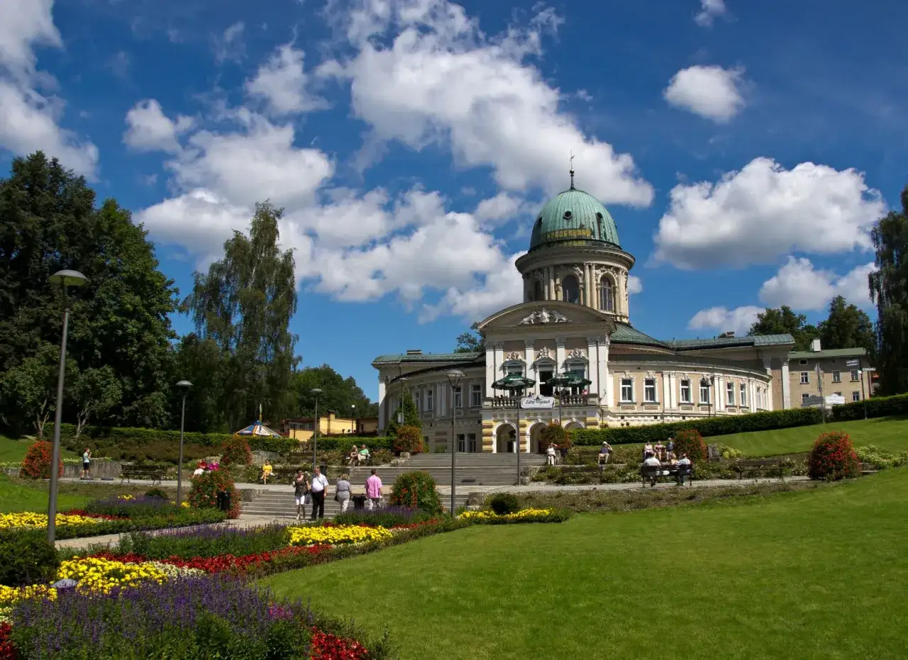 Piękny budynek z zieloną kopułą, otoczony kwitnącym ogrodem i ludźmi spacerującymi po alejkach. To idealne miejsce na relaks, jak w lądku zdroju.
