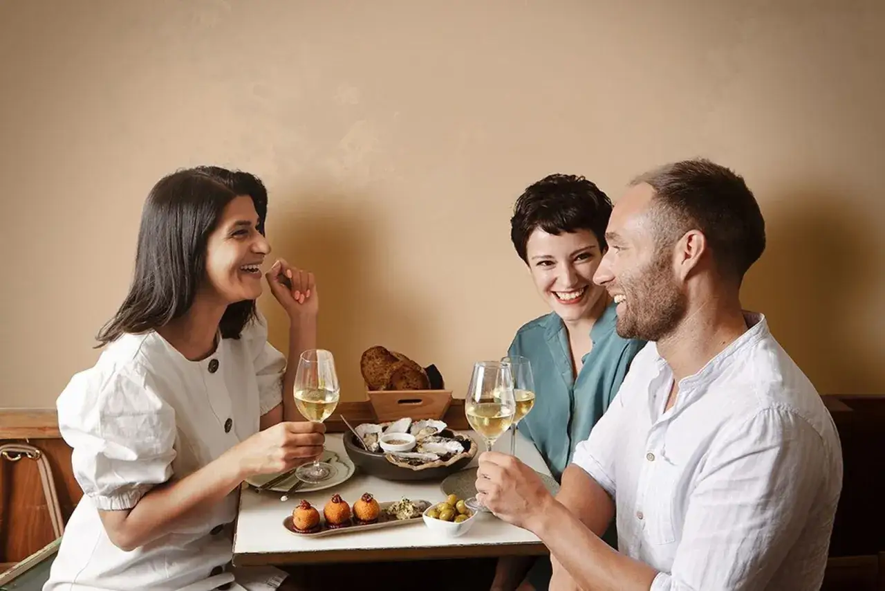 Tres amigos celebran el d&iacute;a del vino con ostras, tapas y copas de vino blanco, riendo y disfrutando de la compa&ntilde;&iacute;a.