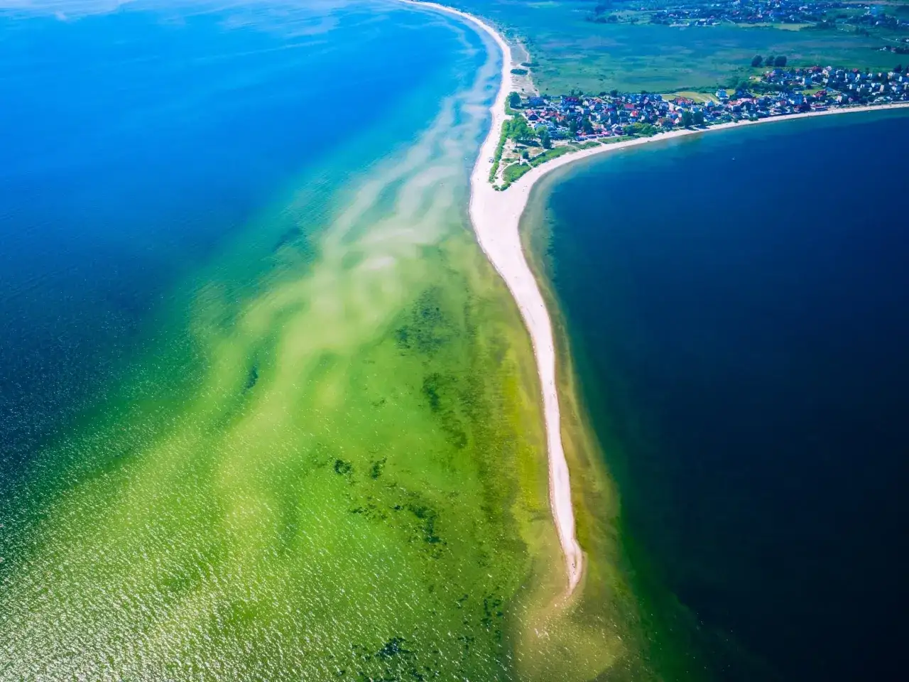 Wąska plaża na cyplu helskim, otoczona turkusową i granatową wodą. W oddali widać zabudowania.