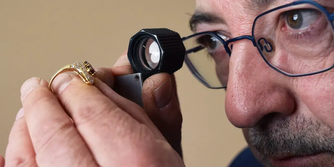 Un homme avec des lunettes examine une bague en or sertie de diamants et d'une pierre rouge &agrave; l'aide d'une loupe, pour faire estimer un bijou.