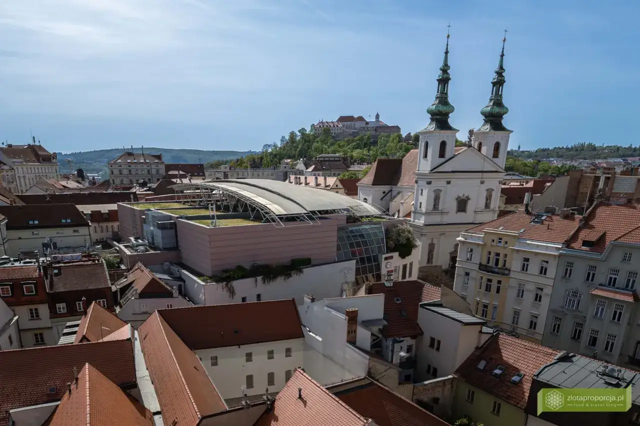 Widok na dachy Brna, z kościołem o dw&oacute;ch wieżach i zamkiem na wzg&oacute;rzu. Wśr&oacute;d miejskich atrakcji wyr&oacute;żnia się nowoczesny budynek z zielonym dachem.
