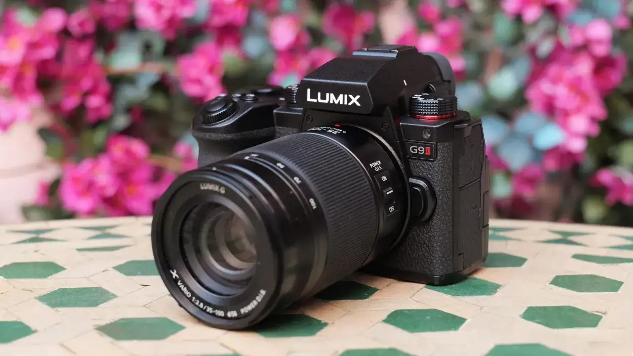 Nowy aparat Lumix G9 II z obiektywem na stoliku z mozaiką, w tle kwitnące różowe kwiaty.