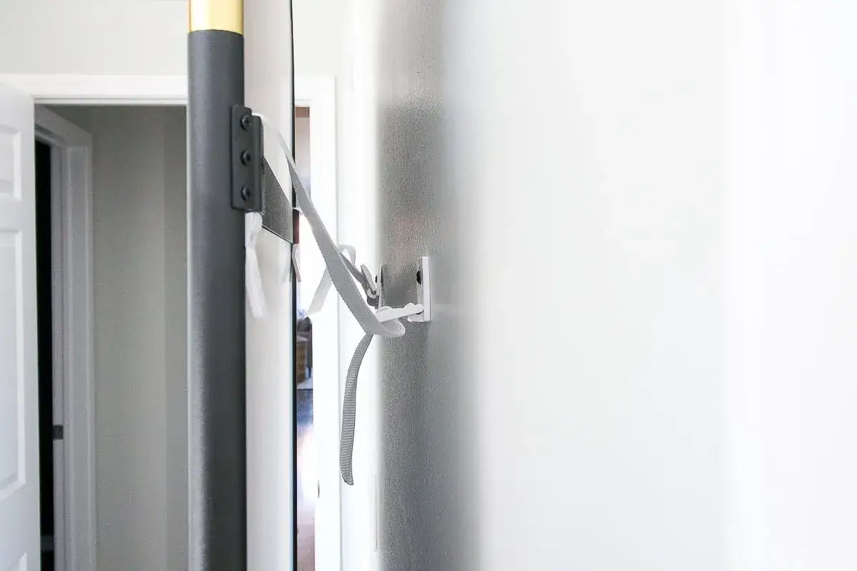 Sistema de anclaje para como fijar un espejo apoyado en el suelo. Correa gris sujeta a la pared y al marco del espejo.
