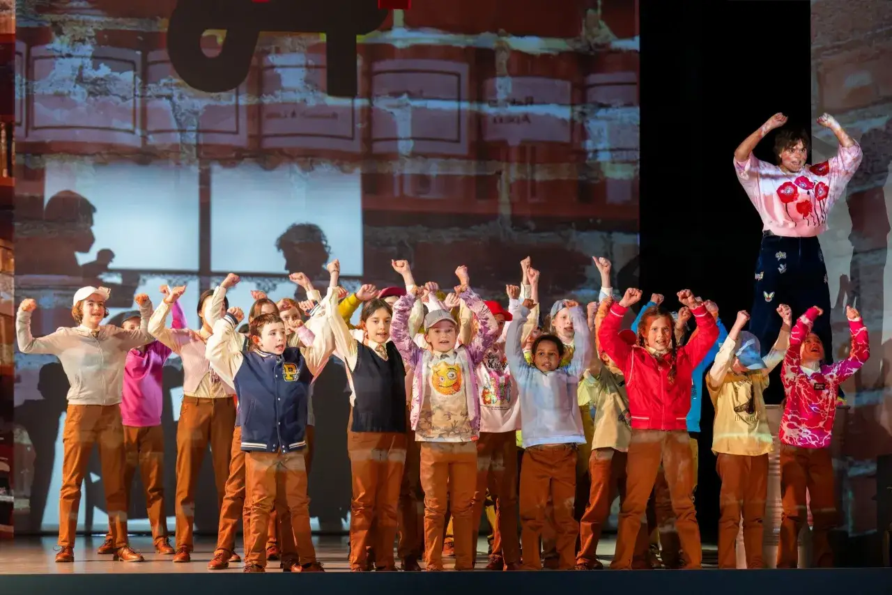 Kinder jubeln auf der B&uuml;hne eines Theaters in Berlin. Eine Frau in einem blumigen Pullover steht &uuml;ber ihnen.