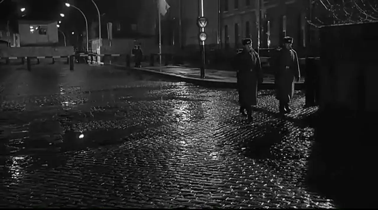 Nocny patrol na mokrym bruku. Dwóch żołnierzy w płaszczach przemierza pustą ulicę, jakby wyszli prosto z zimnej strefy.